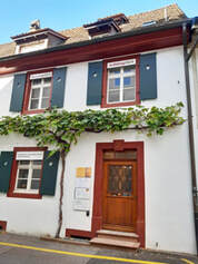 Eingang von Strasse zur Praxis Riehen Dorf. Historisches Gebäude mit Efeu begrünt, dunkel braunrot und weiss. Schöne antike Holztüre.